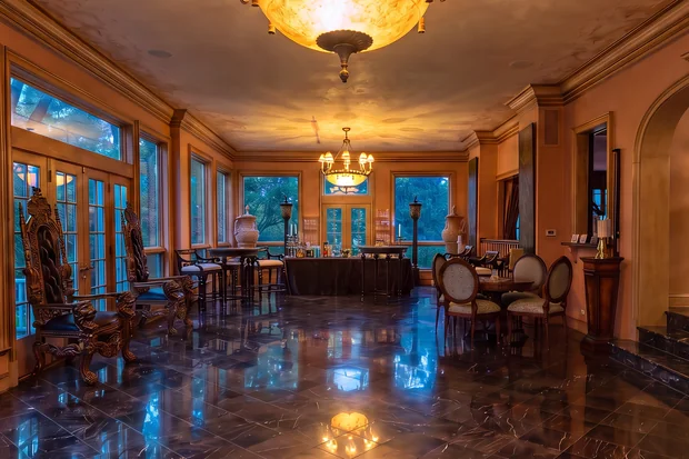 The ballroom at dusk: a lit ceiling dome and chandelier, tall windows, a bar at the far end, and a dark marble floor.